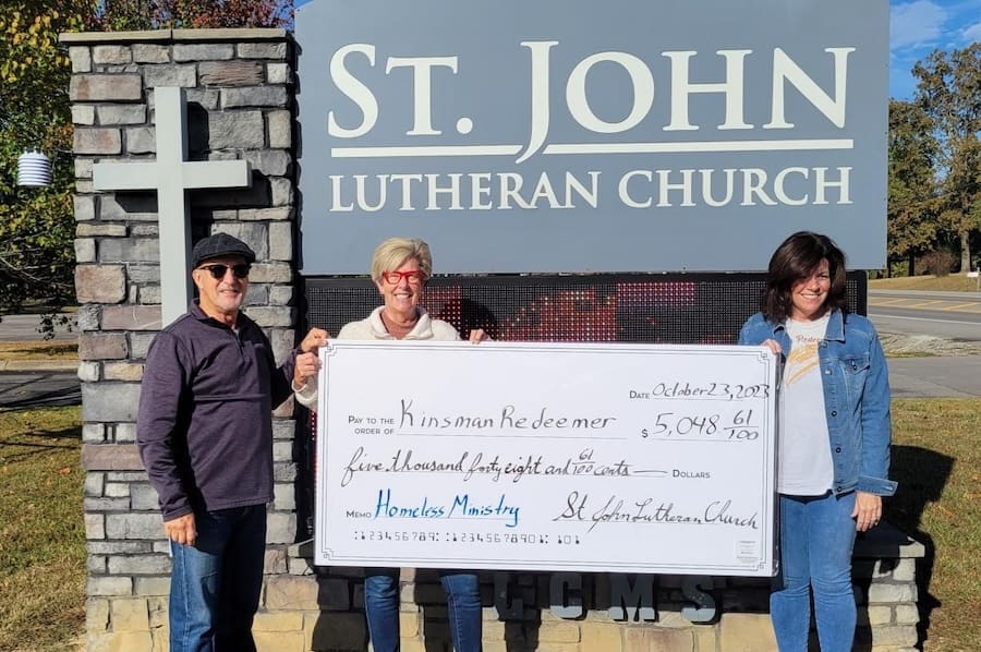 Three people stand in front of a St. John Lutheran Church sign, holding a large donation check made out to Kinsman Redeemer for $5,048.61 in support of Homeless Ministry. Kinsman Redeemer Homeless Ministry in Dickson County Tennessee